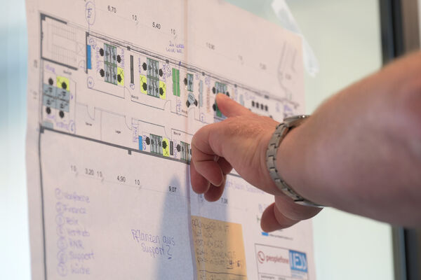 Hand zeigt auf einen Papierplan an einer Glaswand.
