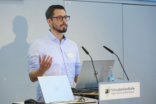 Eine Person steht auf der Bühne mit Mikrofon und hält einen Vortrag.