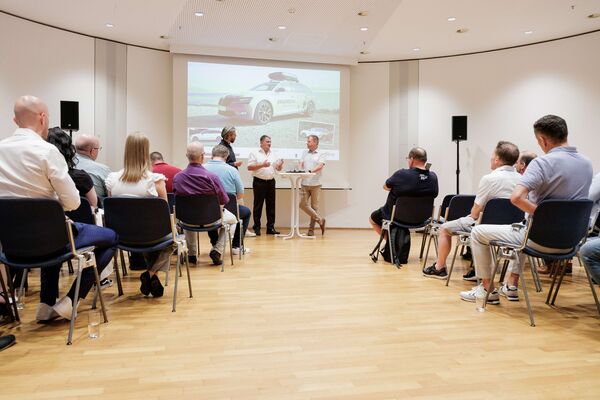 Drei Personen stehen vor einer Leinwand mit Headset und führen ein Gespräch. Der Blick auf die Personen ist aus dem Zuschauerraum.