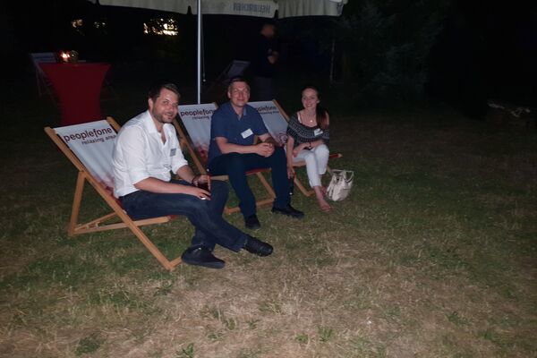 Drei Personen im Liegestuhl schauen in die Kamera. Die Liegestühle stehen auf einer Wiese.