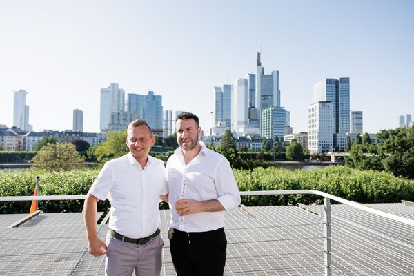 Zwei Personen stehen auf einer Dachterrasse. Im Hintergrund ist die Skyline von Frankfurt zu sehen.
