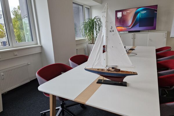 Segelschiff auf Konferenztisch. Im Hintergrund ist ein Bildschirm, auf der linken Seite sind Bürofenster.