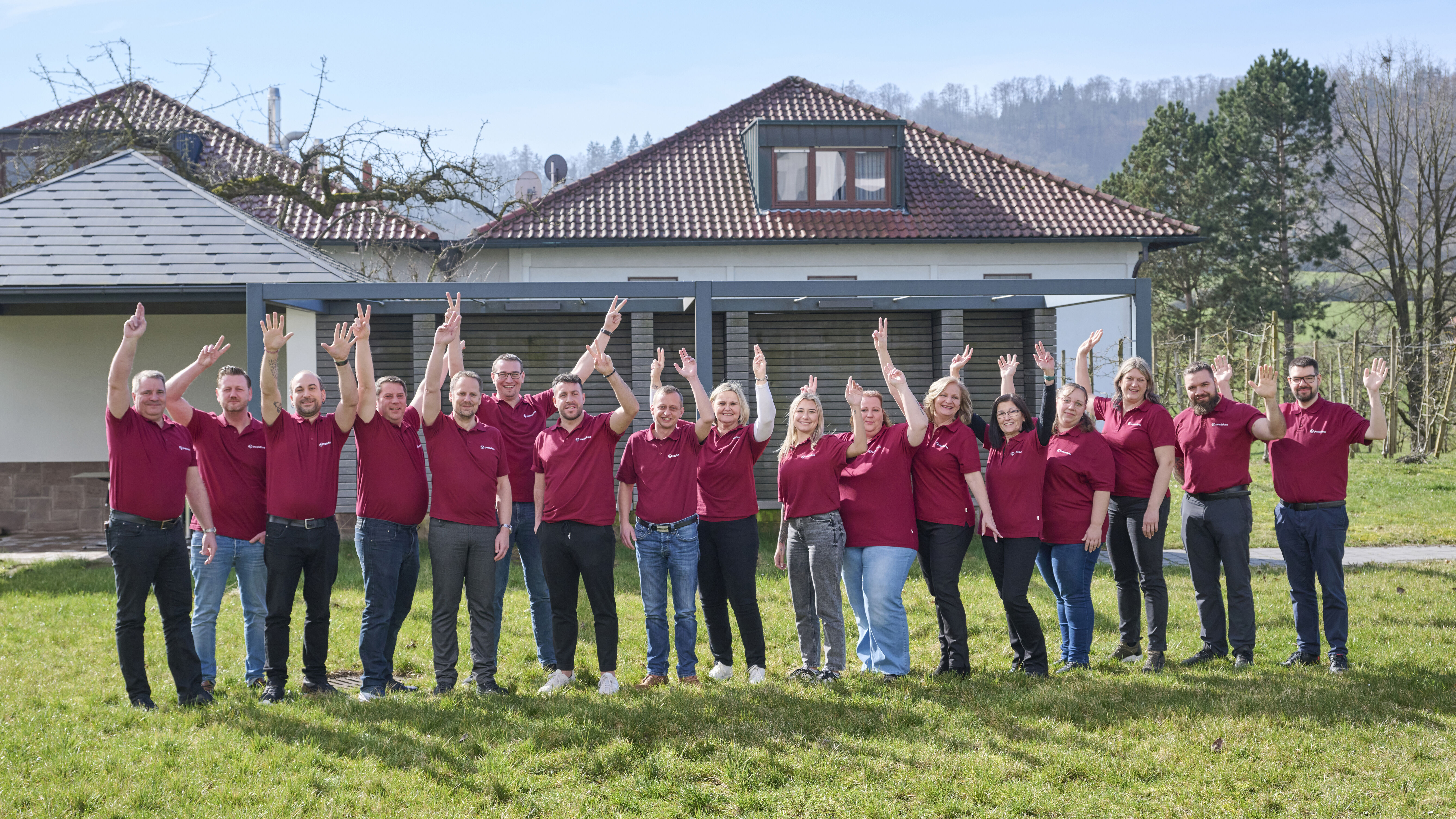 peoplefone Mitarbeiter stehen auf dem Rasen vor einem Geäbude und winken in die Kamera. Alle tragen ein rotes Polo Shirt.
