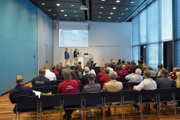 Zuschauer sitzen im Raum und hören einem Vortrag zu. Auf der Bühne stehen zwei Personen und halten einen Vortrag.