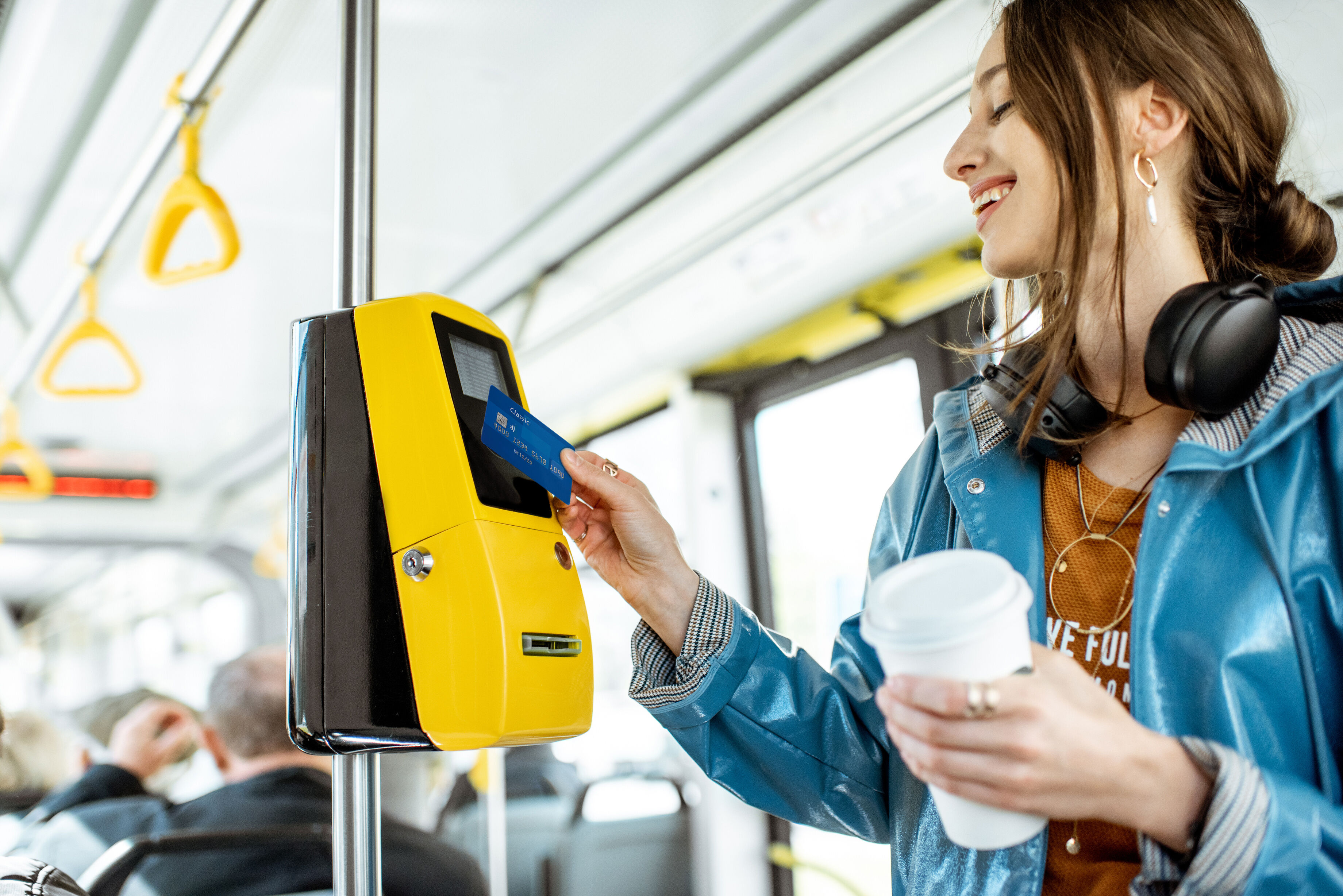 Eine lächelnde Frau steht im Bus. Sie hält in der linken Hand einen Coffe-to-go Becher und mit der rechten Hand eine Karte an den Fahrkartenautomaten.