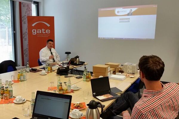 Personen am Tisch sitzend mit Laptops schauen auf eine Bildschirmleinwand.