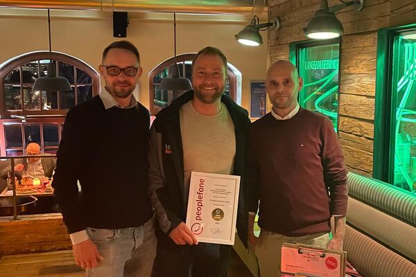 Drei Personen schauen in die Kamera und halten eine Urkunde in einem Lokal.