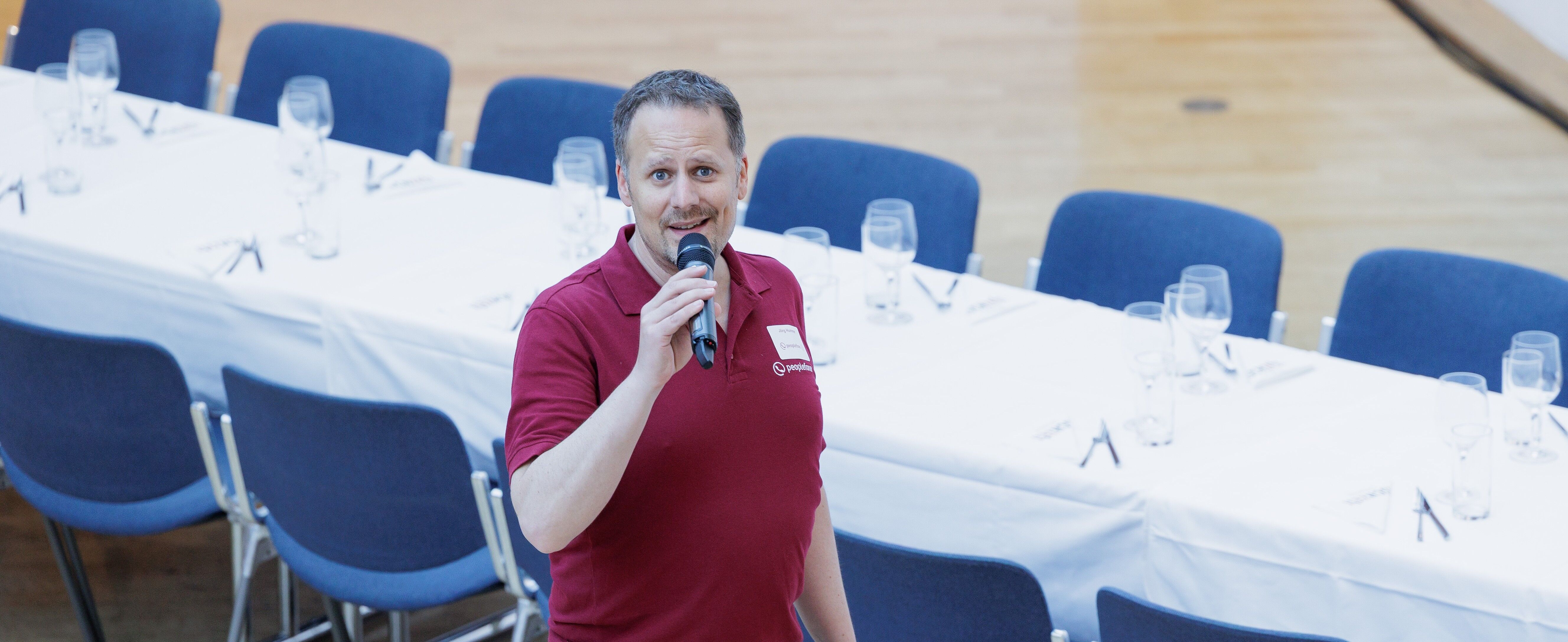 Männliche Person steht mit einem Mikrofon in der Hand vor einem gedeckten Tisch mit Bestuhlung.