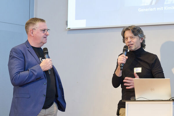 Zwei Personen stehen auf der Bühne mit Mikrofon und halten einen Vortrag.