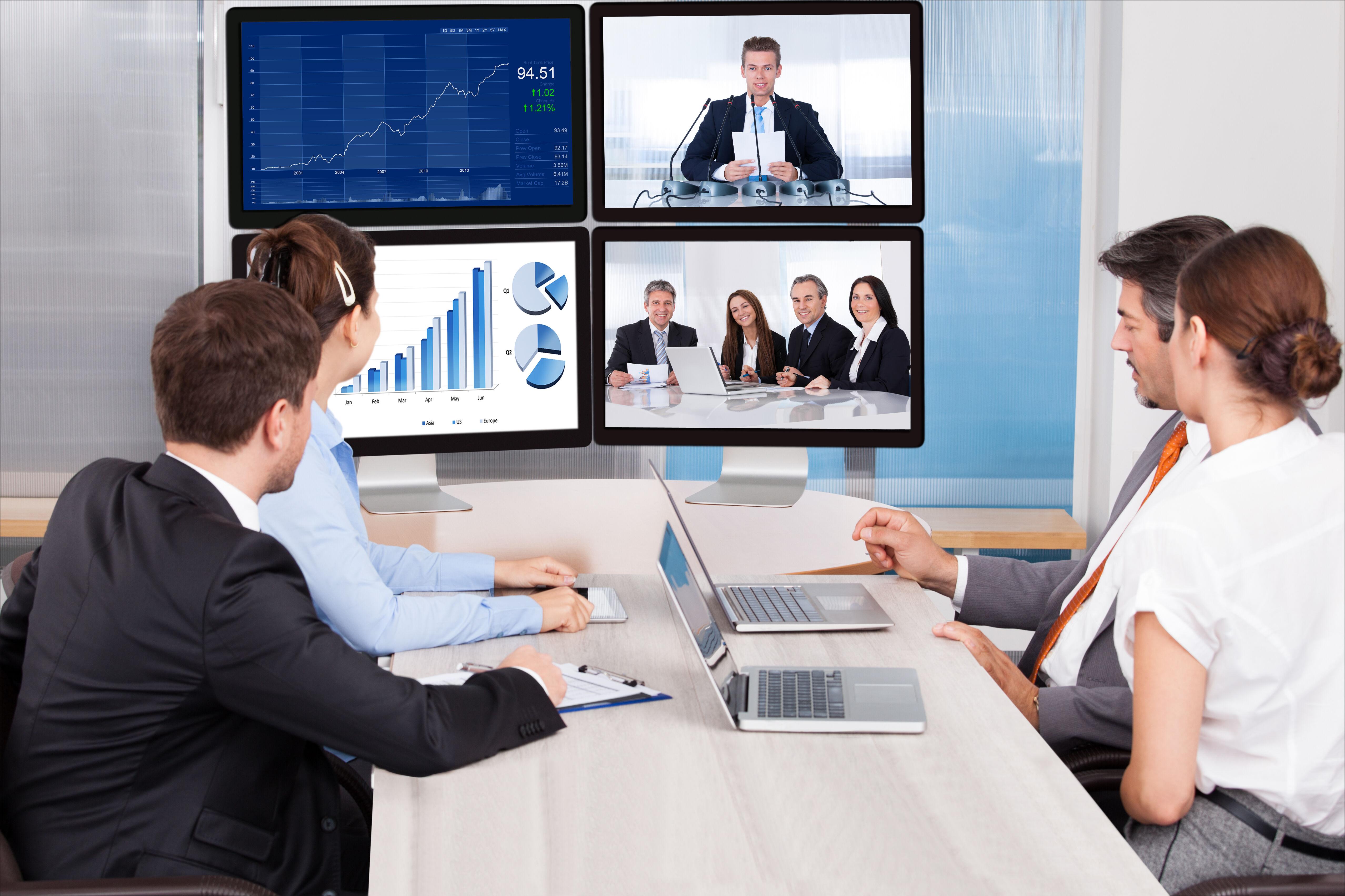 Vier Personen sitzen an einem Konferenztisch und schauen auf vier Bildschirme. Dort sind Grafiken und weitere Personen aus einer Videokonferenz sichtbar. Vor ihnen stehen zwei Laptops.