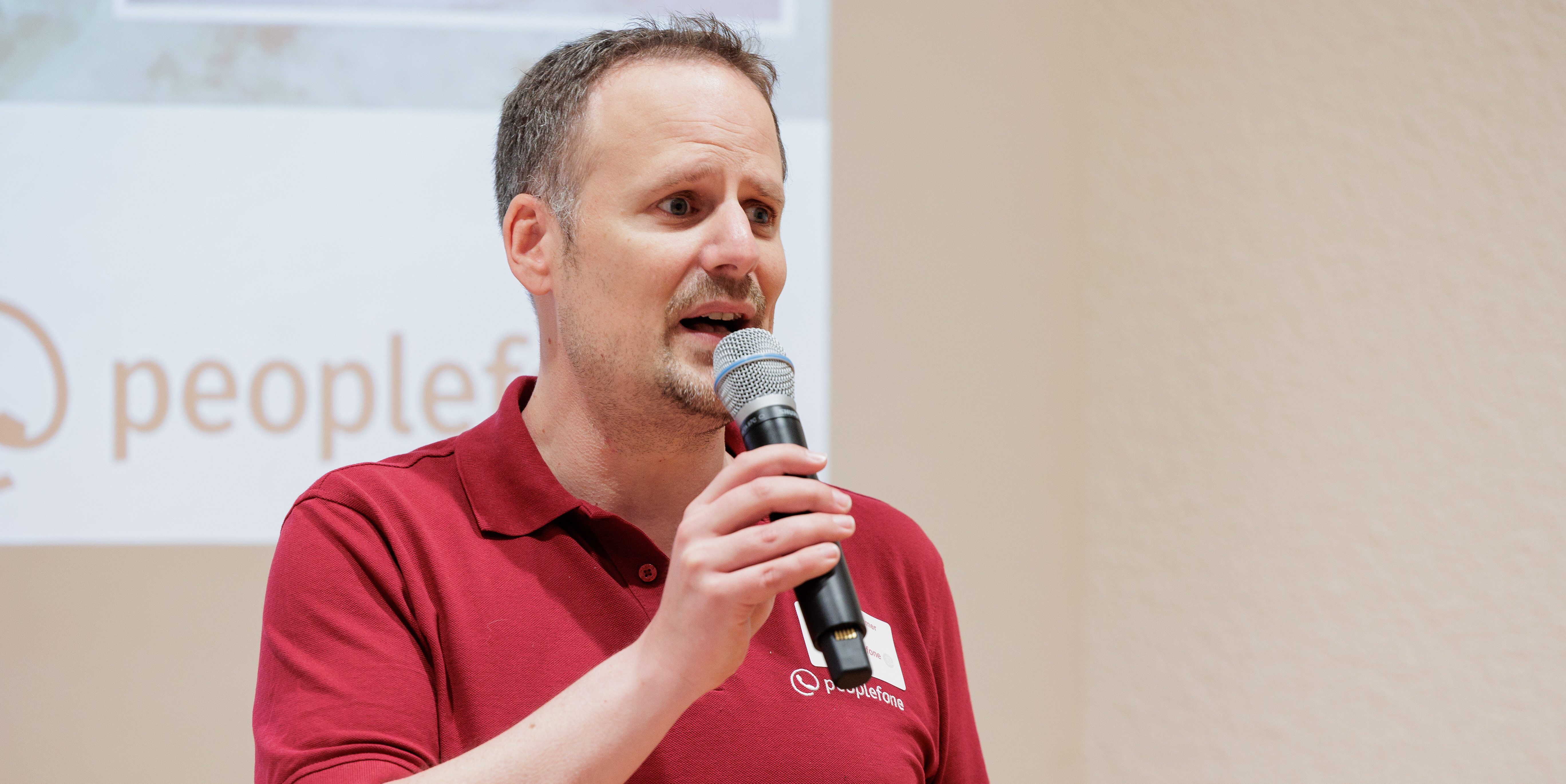 Männliche Person steht mit einem Mikrofon in der Hand vor einer Leinwand und begrüßt die Gäste.