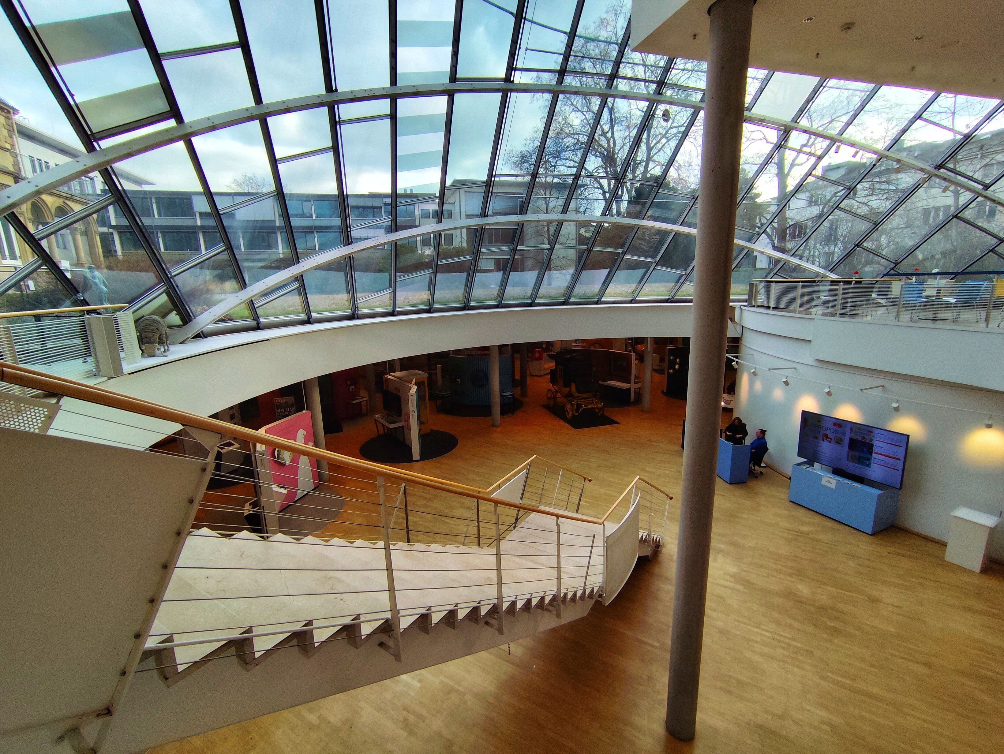 Treppe führt in einen Lichthof mit Glaskuppel und Blick auf mehrere Gebäude.
