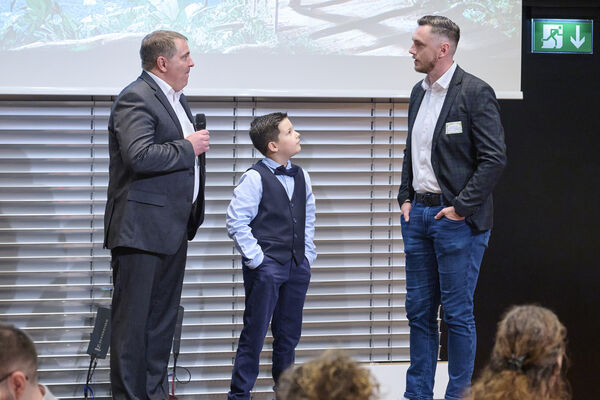 Drei Personen stehen auf einer Bühne und führen ein Interview. Im Hintergrund ist ein Spendenscheck eingeblendet auf einer Leinwand.