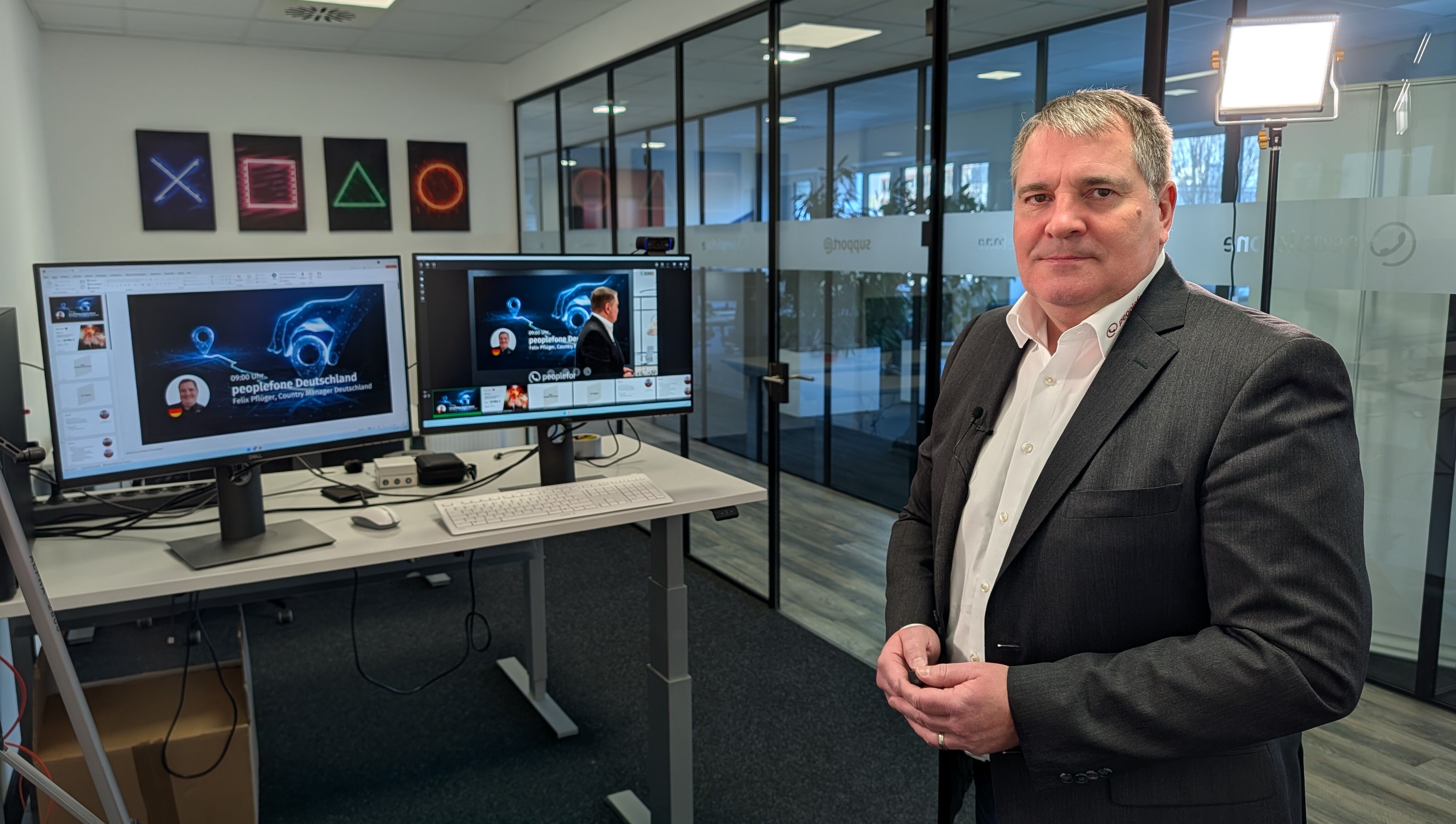 Referent steht im Studio und schaut in die Kamera. Im Hintergrund wird auf Bildschirmen ein Webinar übertragen.