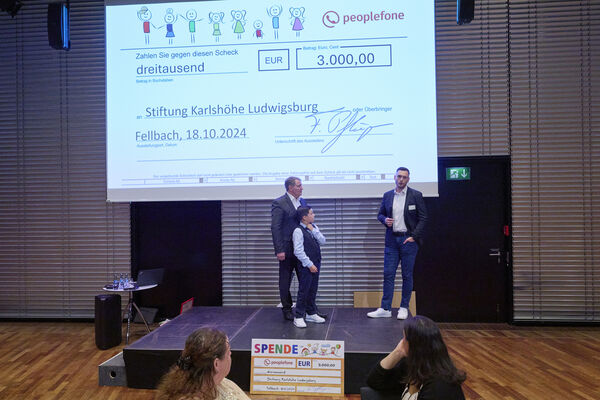 Drei Personen stehen auf einer Bühne und führen ein Interview. Im Hintergrund ist ein Spendenscheck eingeblendet auf einer Leinwand.