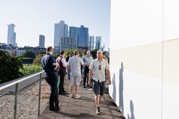 Mehrere Personen stehen auf einer Dachterrasse. Im Hintergrund sieht man die Frankfurter Skyline.