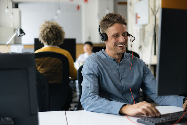 Männliche Person sitzt mit einem Headset vor einem PC in einem Office.