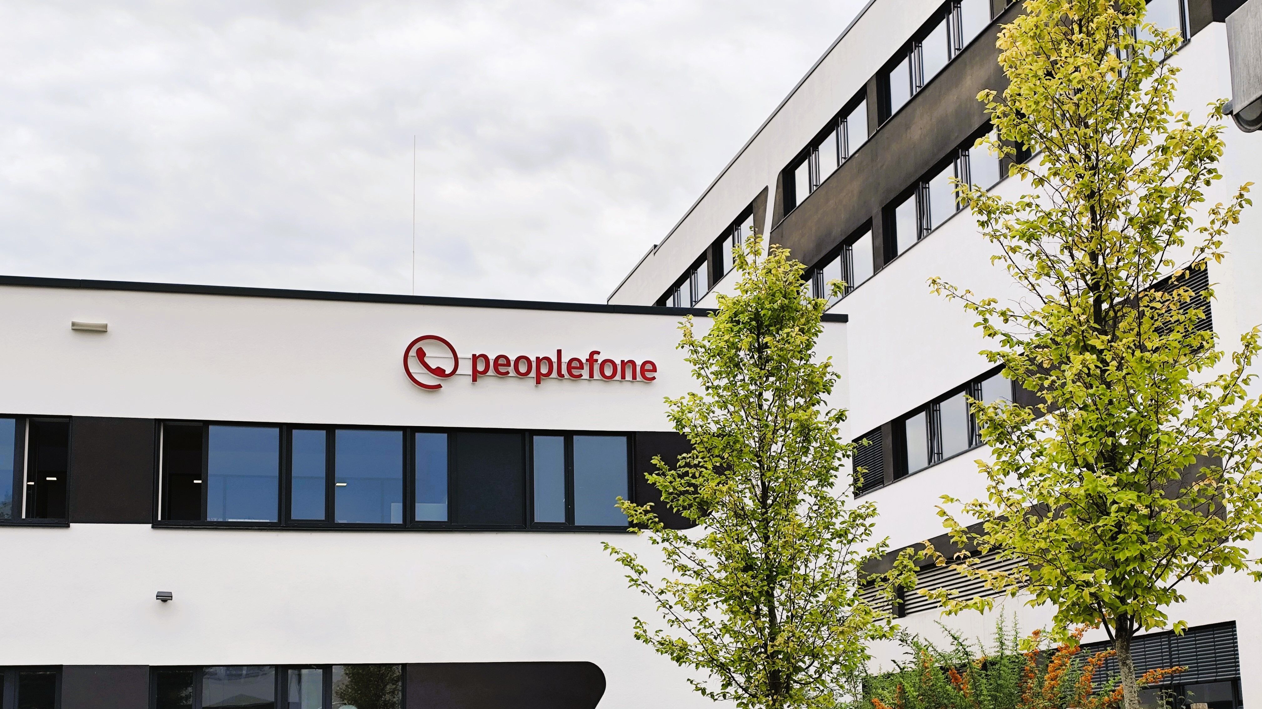 Zwei weiße Firmengebäude mit Flachdach stehen nebeneinander. Davor ist ein grüner Baum und das Logo der peoplefone ist am ersten Gebäude angebracht.