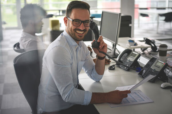 Männliche Person sitzend am Schreibtisch telefoniert und schaut in die Kamera.