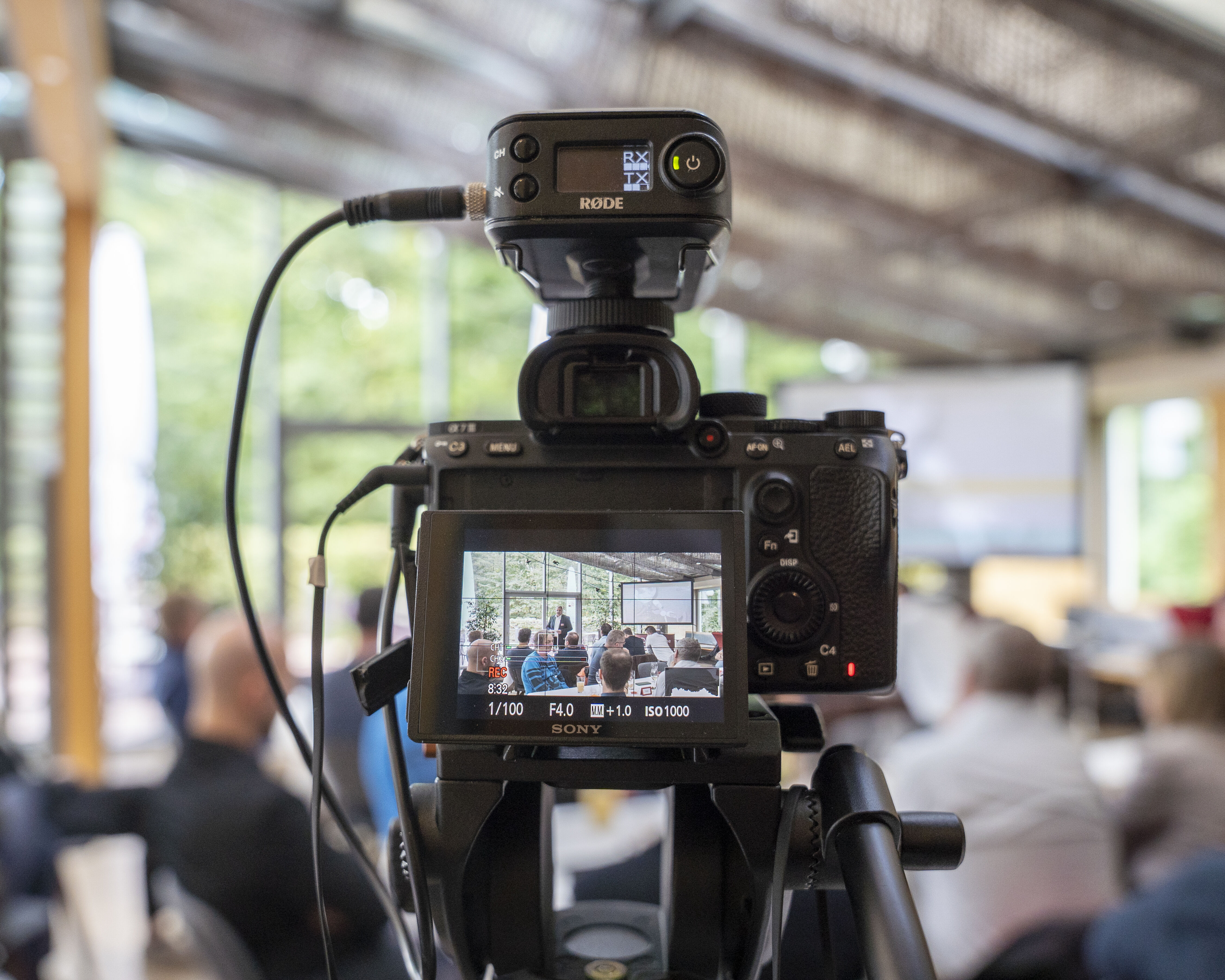 Blick durch eine Fotokamera in einen Raum mit sitzenden Menschen, die einem Referenten zuhören.