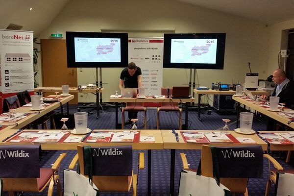 Tische und Stühle in U-Form in einem Raum. Auf der Vorderseite zwei Bildschirme und ein Referent am Laptop.