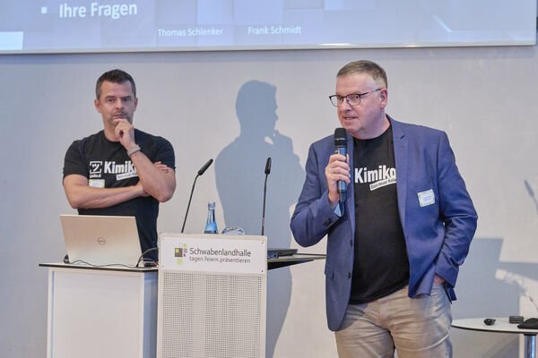 Zwei Personen stehen auf der Bühne mit Mikrofon und halten einen Vortrag.